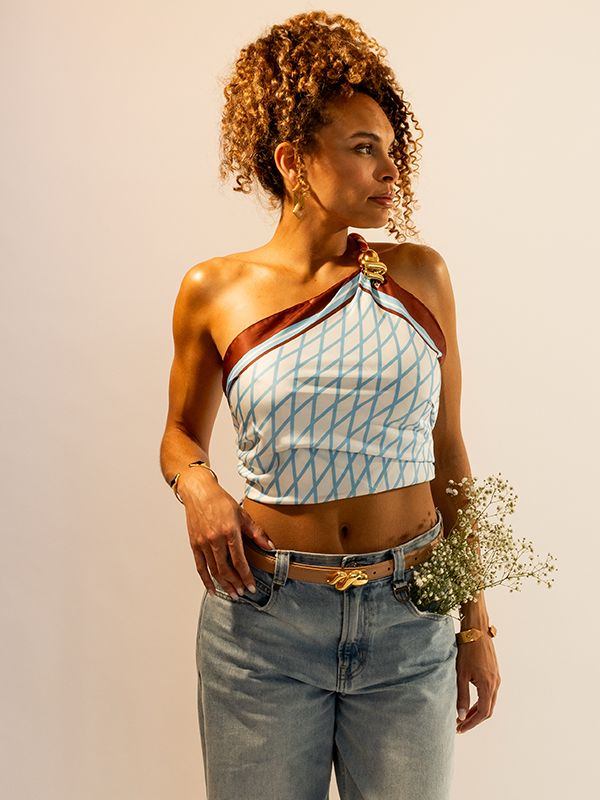 A woman modeling a one-shoulder crop top with a light blue and white geometric RUAHH scarf, Snake belt brown trim, and a gold decorative clasp, paired with light-wash blue jeans and holding a small bouquet of baby's breath against a plain beige studio background.