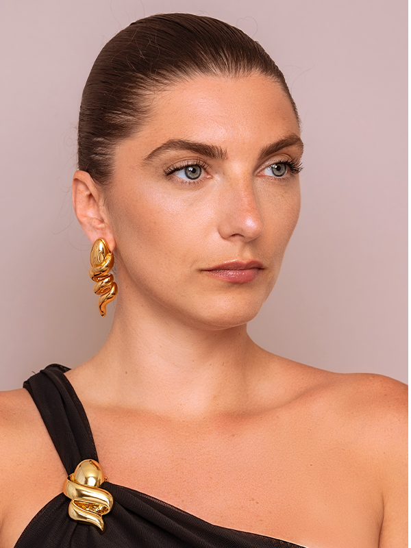 A close-up portrait of a woman wearing a sculptural, twisted gold earring and a matching gold snake-braided shoulder detail on a black one-shoulder top, set against a solid light taupe background.